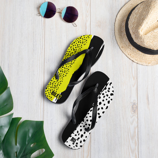 Two pairs of flip-flops with black and yellow, and black and white patterns on a light wooden surface with a hat and sunglasses.
